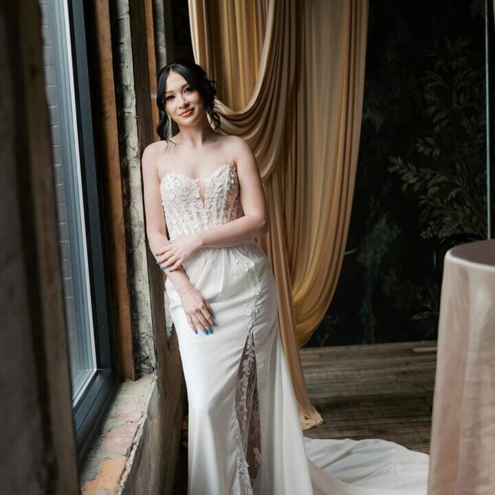 Bride wearing a fitted lace wedding dress standing beside large windows at Atlas Bridal Shop